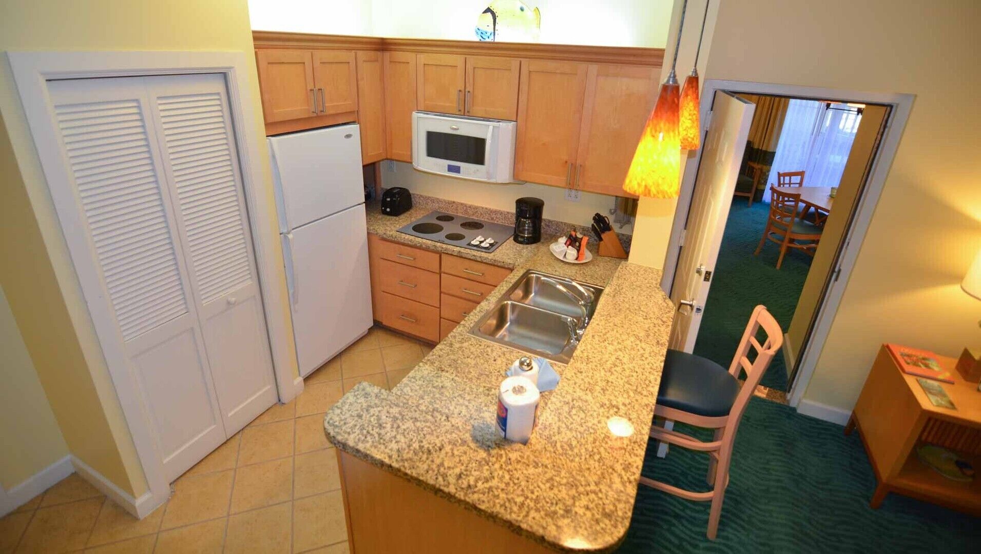 Cocina compacta con refrigerador, microondas, estufa y pileta. Hay una silla y una mesita con una lámpara.