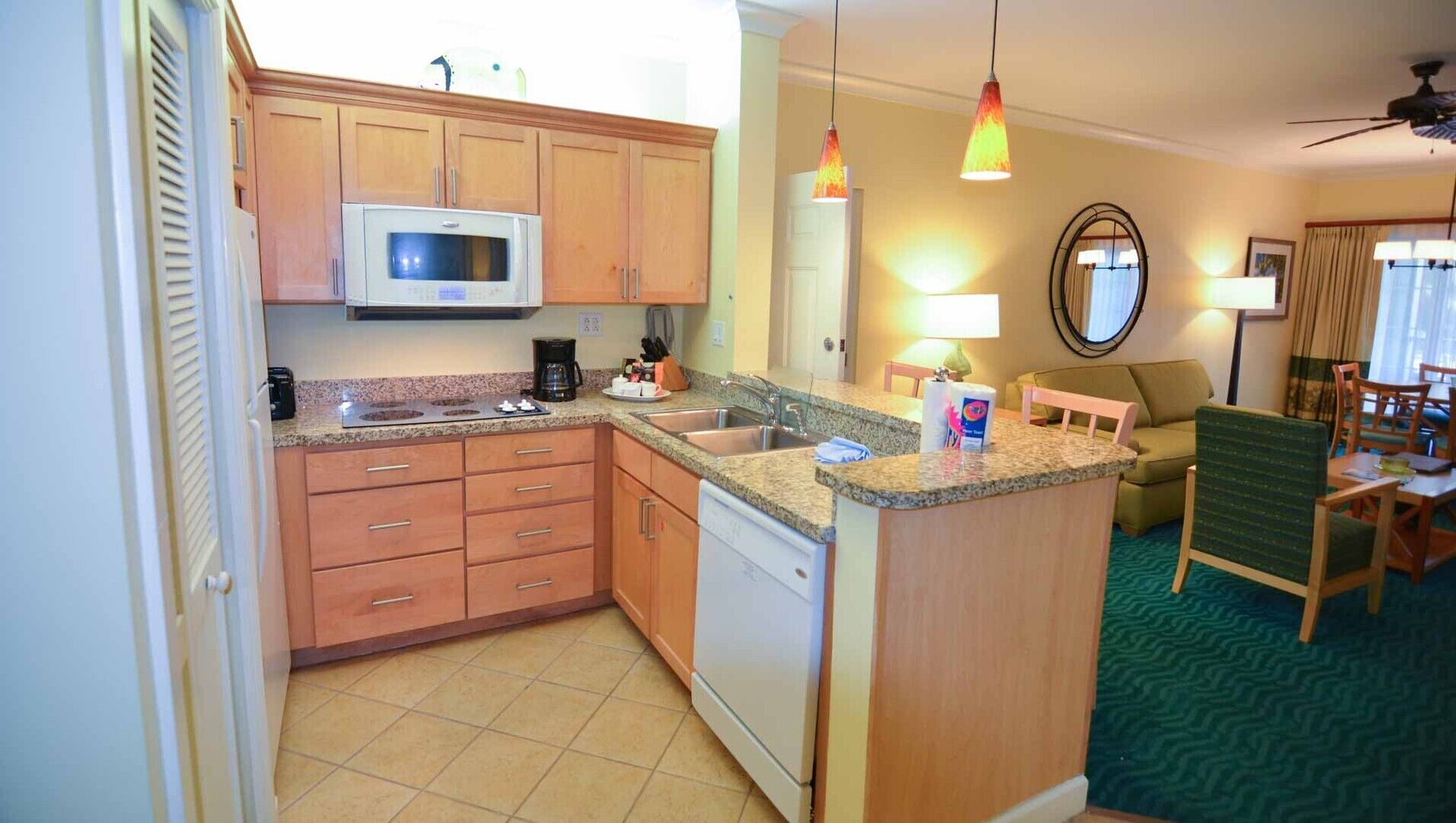 A compact kitchen with wooden cabinets, a microwave, a coffee maker, and a sink. There is a small island with a microwave and a chair. The room has a green carpet and a ceiling fan.