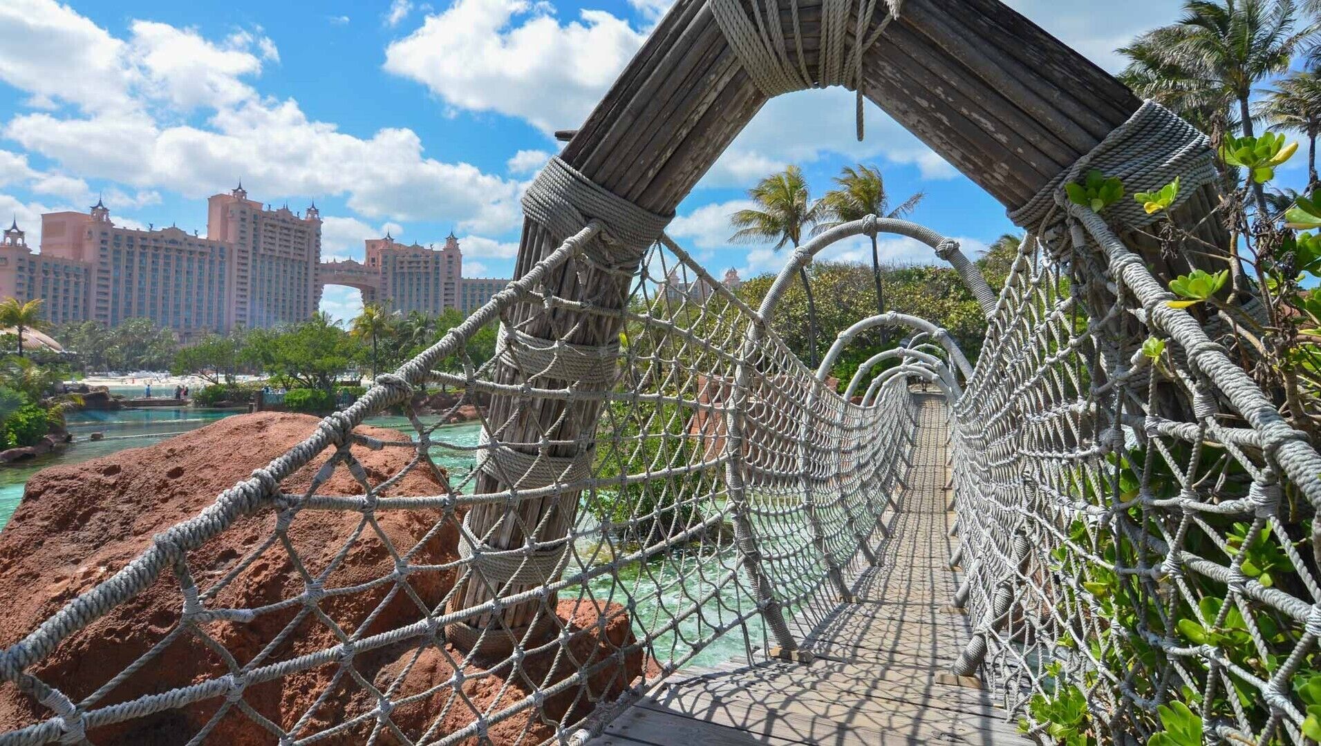 A rope bridge over a body of water with a view of a resort area and high-rise buildings in the background.