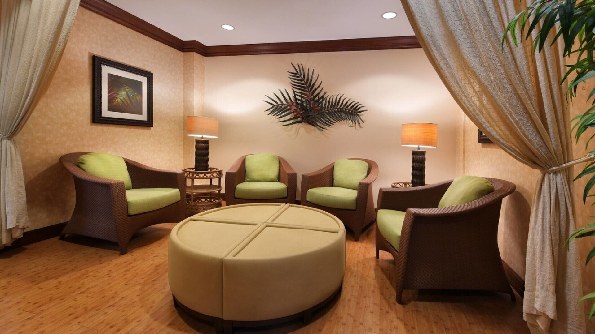 A hotel room with a seating area featuring wicker chairs with green cushions, a round beige ottoman, and two wall-mounted lamps.