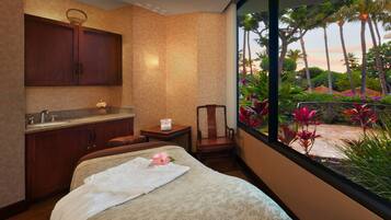 A hotel room with a bed, a bathroom with a sink, a chair, and a view of a garden with palm trees and flowers.