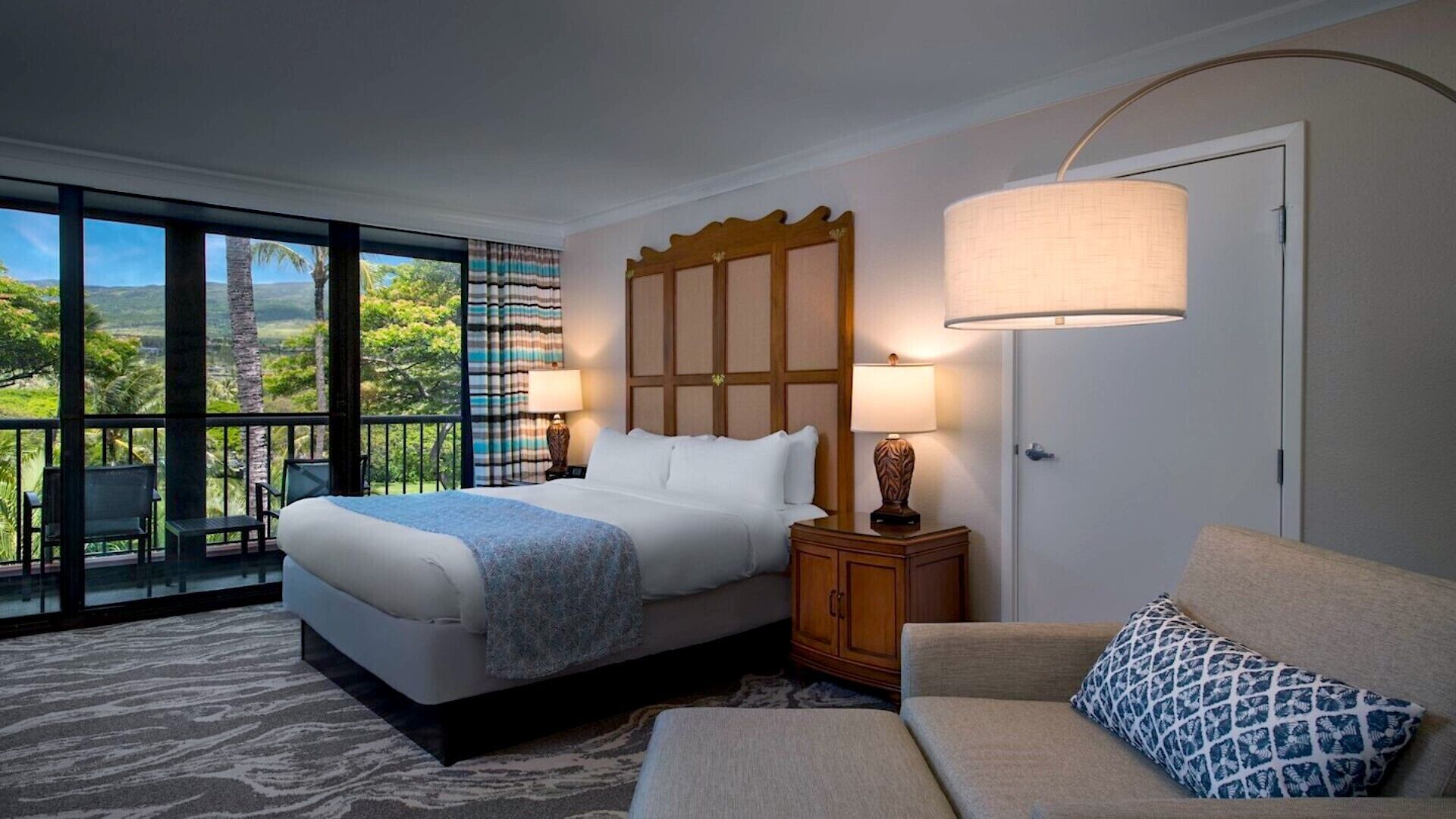 A hotel room with a bed, a nightstand, a lamp, a sofa, and a view of a tropical landscape.