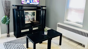 Una sala de estar con un televisor de pantalla plana sobre un soporte negro, un cuadro enmarcado, una mesita con una planta en maceta y un radiador.