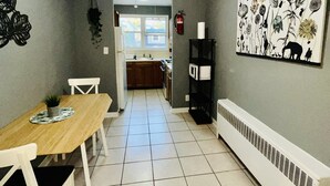 Un comedor con mesa y sillas de madera, suelo de baldosas, cocina al fondo y un radiador fijado a la pared.