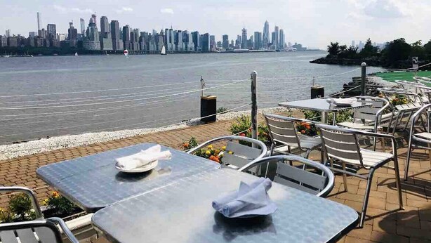 Un área de comedor al aire libre con mesas y sillas, con vista a un río y un horizonte urbano de fondo.