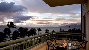 Un balcón con una mesa para dos, con vistas a la costa, palmeras y un atardecer.