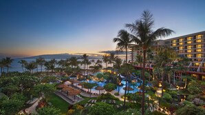 Un resort con un amplio área de piscina, palmeras y vistas al océano al atardecer.