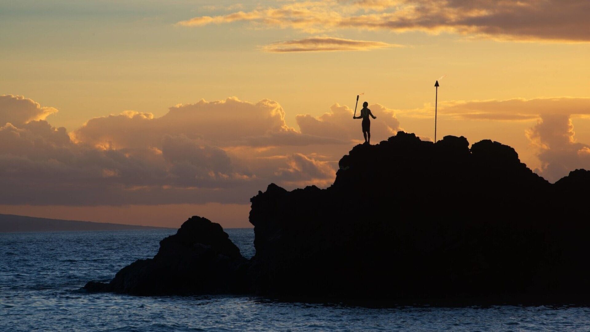 Une personne se tient sur une formation rocheuse, près d’un mât sur lequel flotte un drapeau, et surplombe une étendue d’eau.