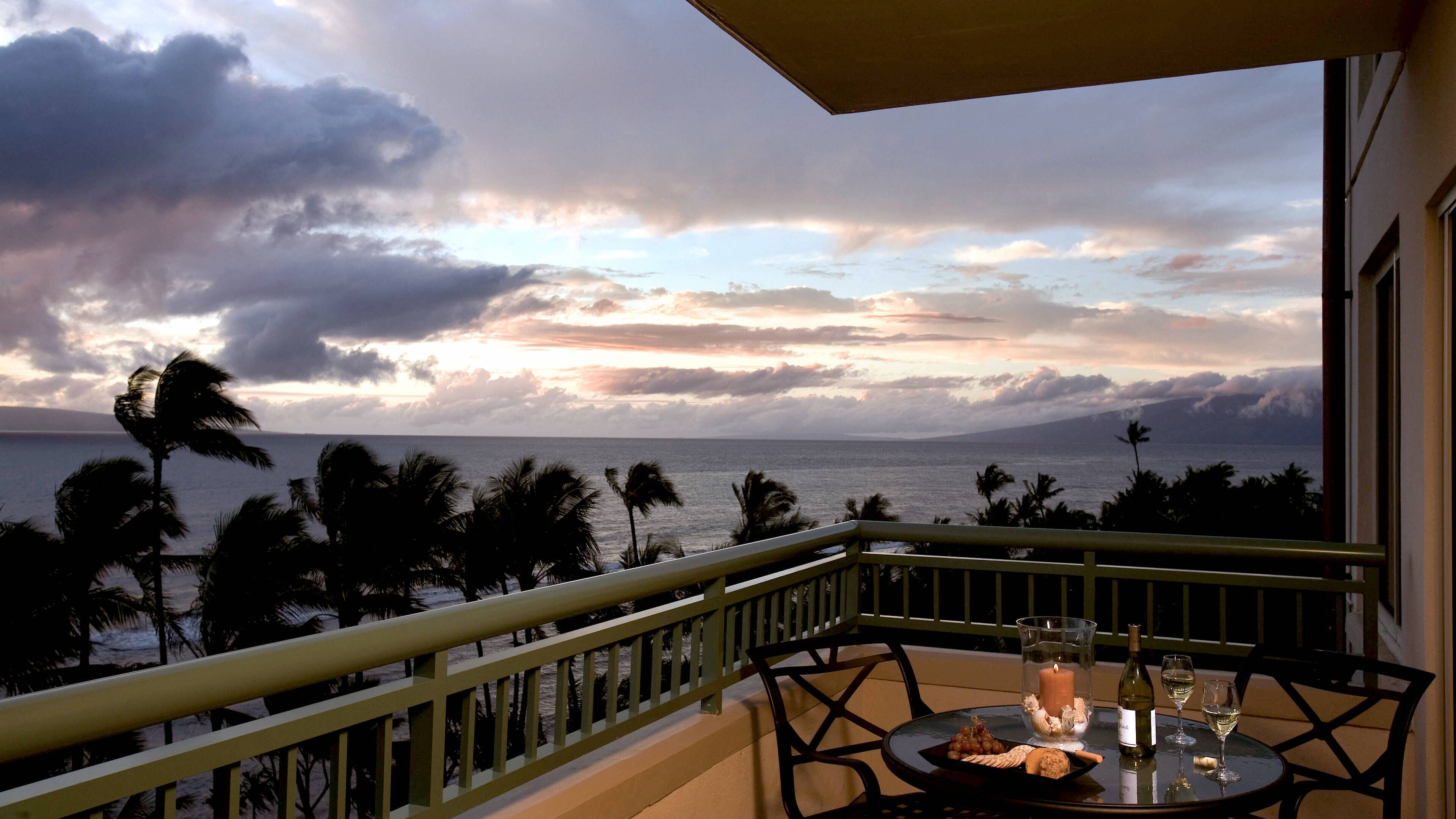 A balcony with a table set for two, overlooking a coastal view with palm trees and a sunset.