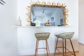 A kitchen with a white countertop, two bar stools, and a decorative mirror with a brick-like frame. - CoolHouses Algarve Luz | Casa Isabell T1 (Luz)