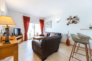 Ein Wohnzimmer mit einer Couch, einem Fernseher, einer Lampe, einem Schachbrett und einem Fenster mit Vorhängen.