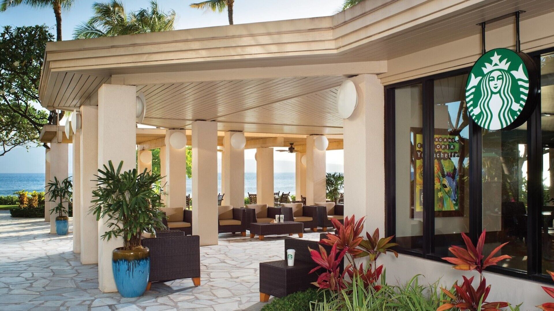 A covered outdoor seating area with wicker furniture and potted plants, a Starbucks sign, and a view of the ocean.