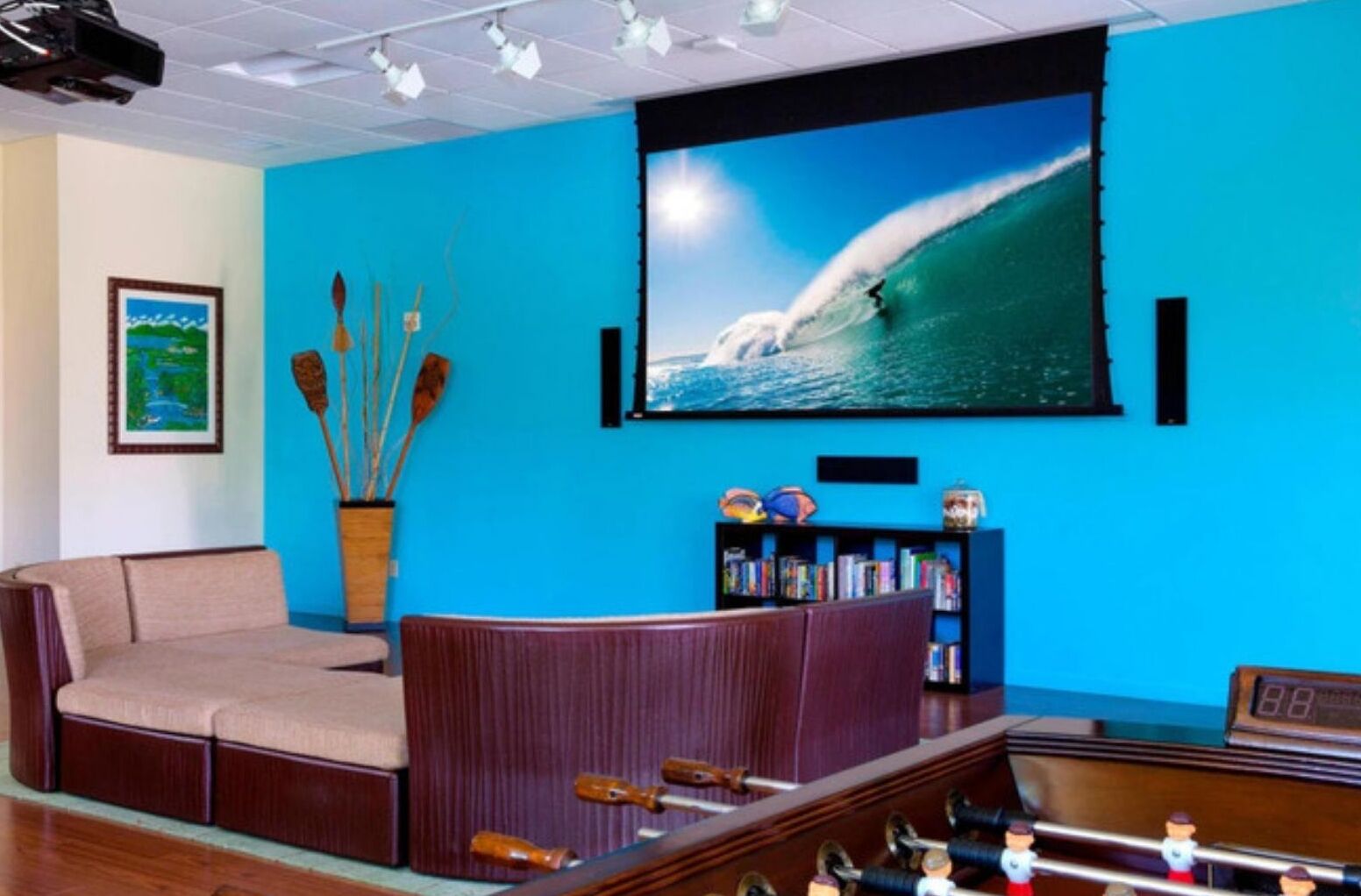 A room with a large flat-screen TV mounted on a blue wall, a wooden coffee table, a bookshelf, and a small table with a clock.