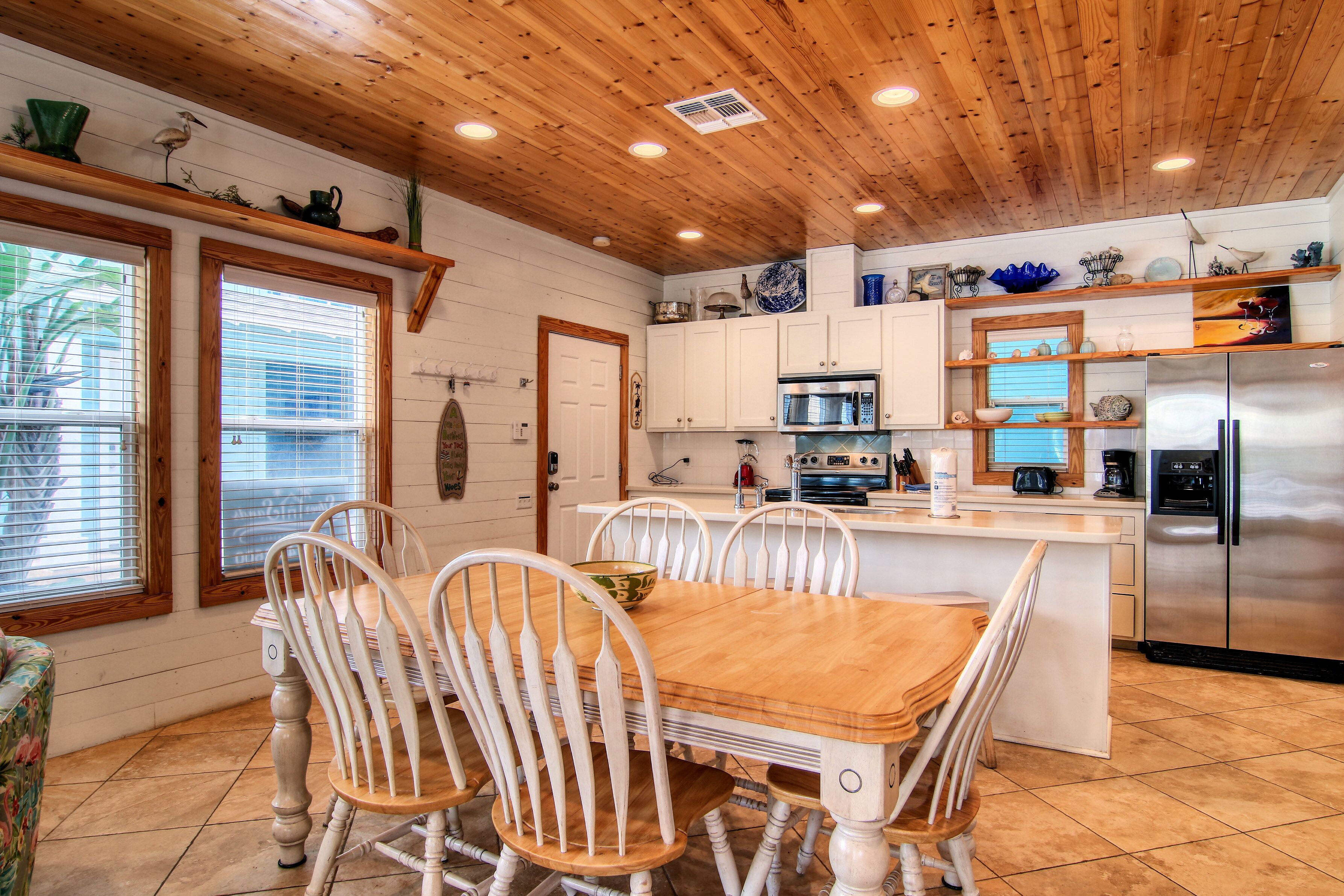 A kitchen with wooden ceiling, white cabinets, stainless steel appliances, and a dining area with white chairs and a wooden table.