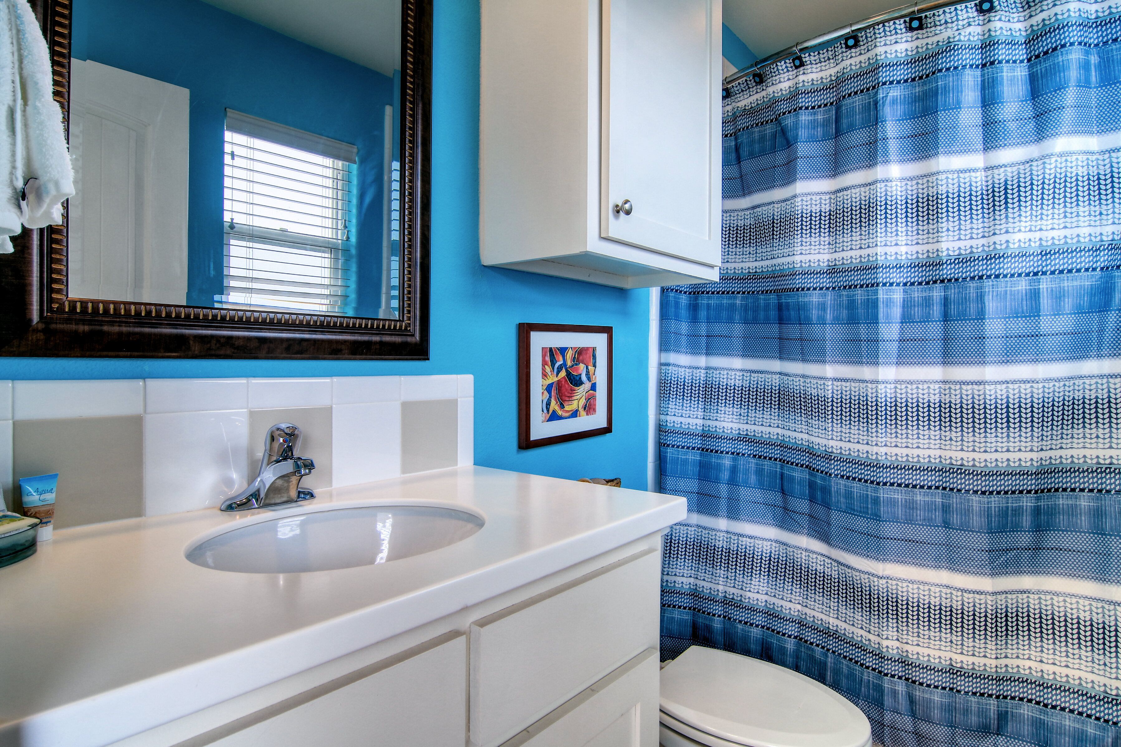 Une salle de bain avec un lavabo blanc, un miroir, un tableau encadré au mur et un rideau de douche à rayures bleues.