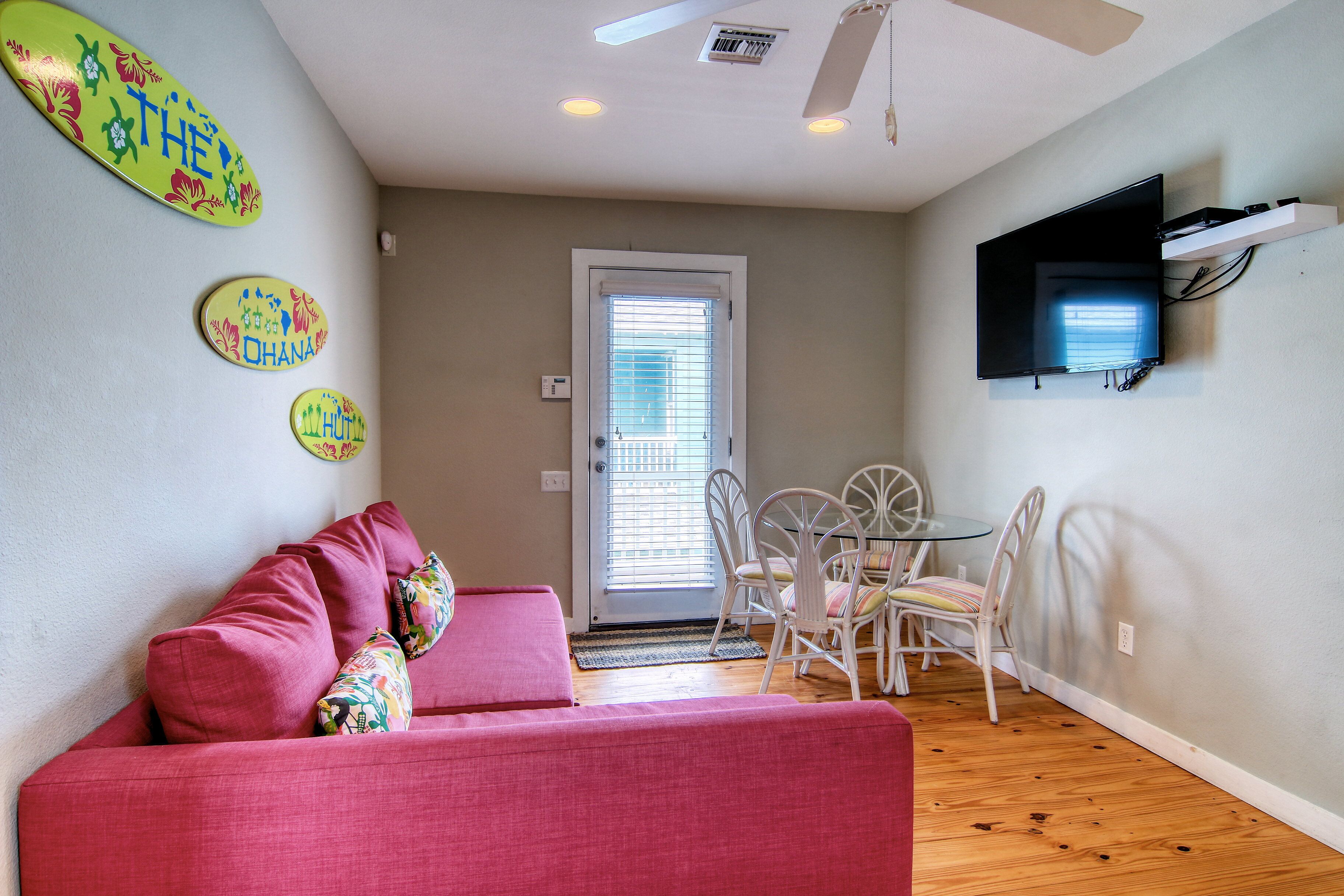 A living room with a pink sofa, a dining table with chairs, a TV mounted on the wall, and a door with blinds.