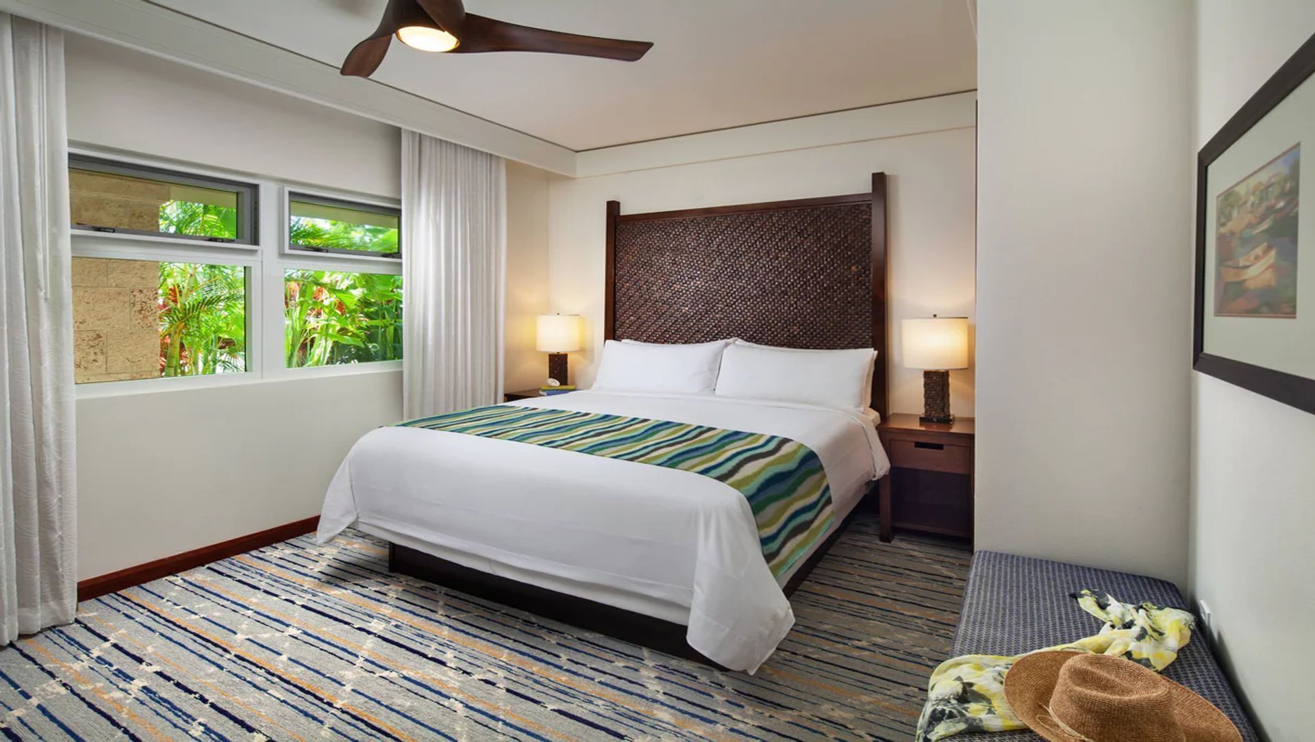 A hotel room with a bed, a ceiling fan, a window with curtains, a wall-mounted TV, and a framed picture.
