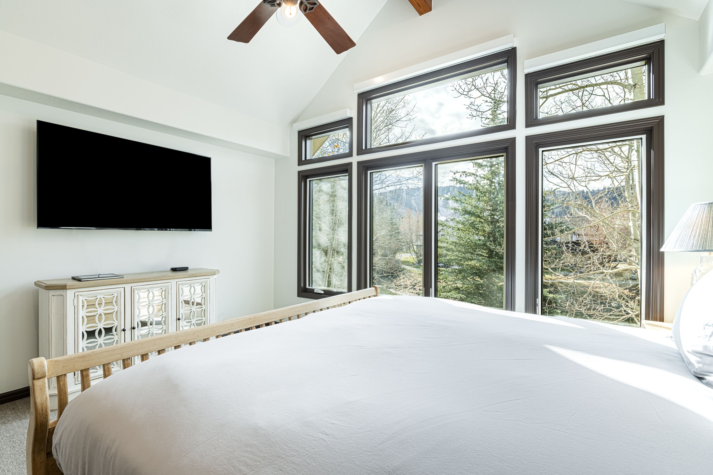 Un dormitorio con una cama grande, un televisor montado en la pared y varias ventanas con vistas a árboles y montañas.