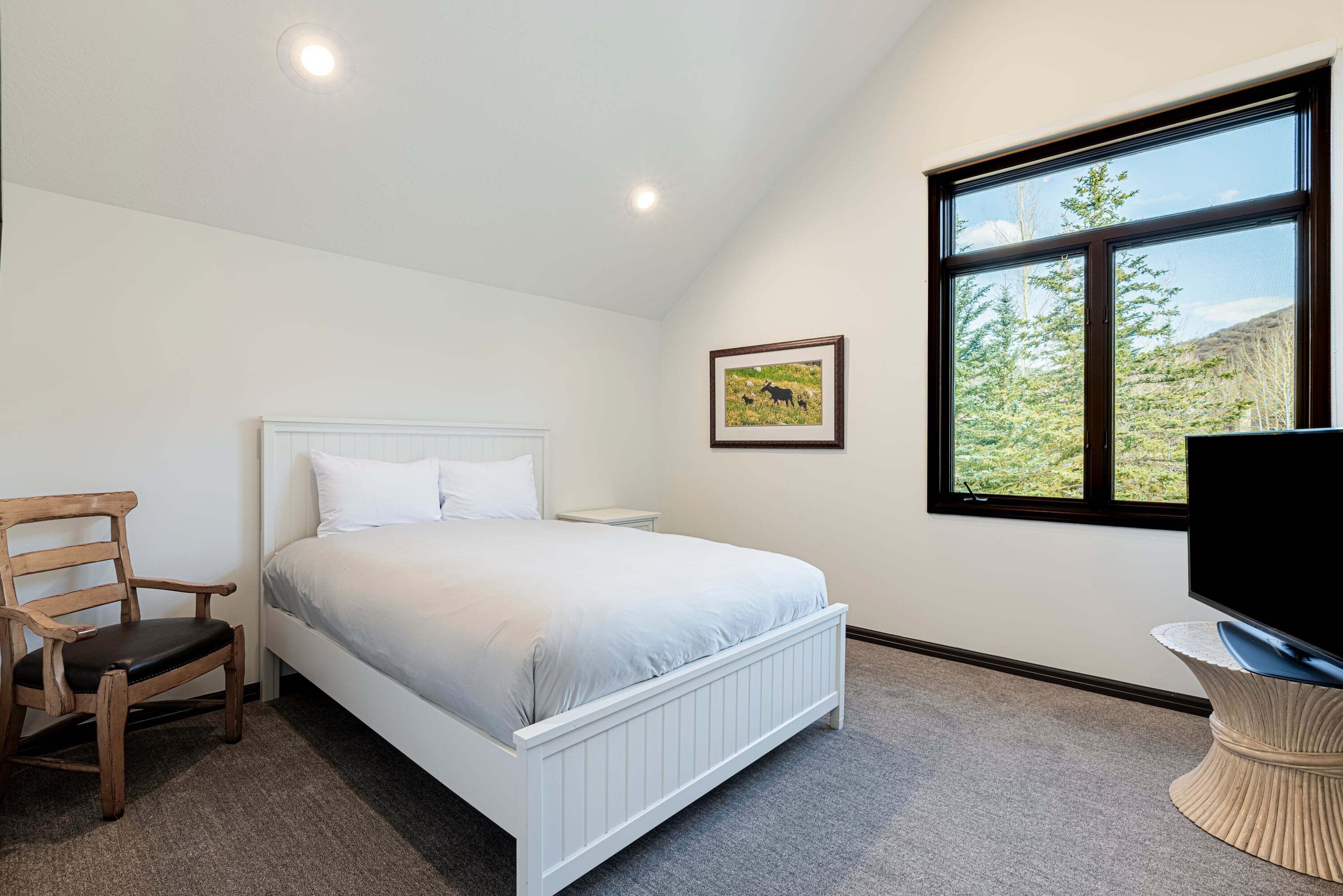 A bedroom with a bed, a chair, a bedside table, a TV, a window, and a framed picture on the wall.