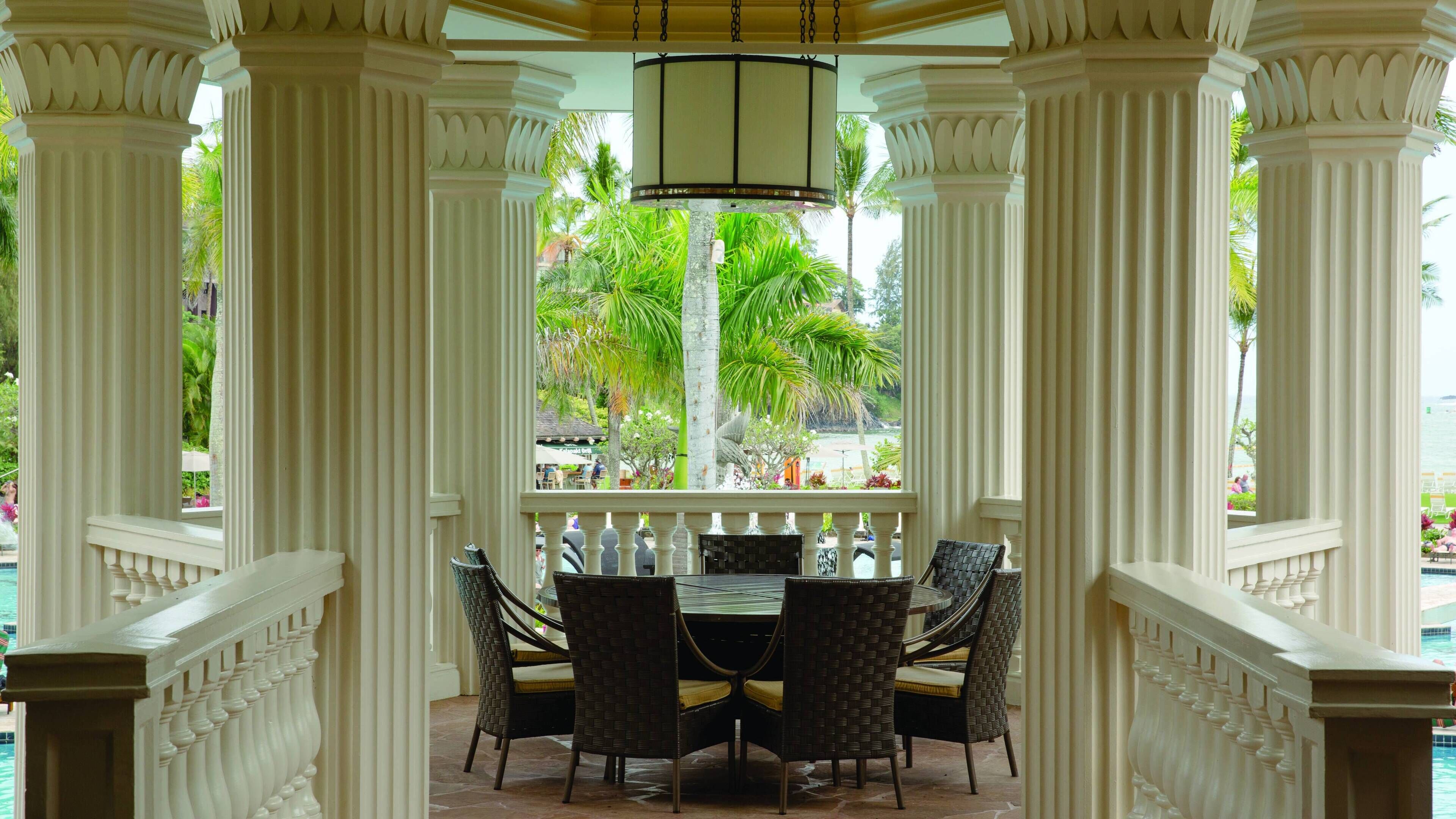 A covered outdoor seating area with a table and chairs, surrounded by white columns and a view of the pool and palm trees.