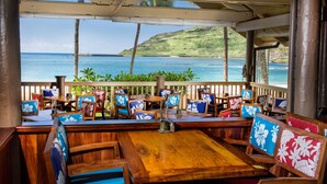 Un restaurante frente al mar con mesas y sillas de madera, vista al océano y una terraza de madera.