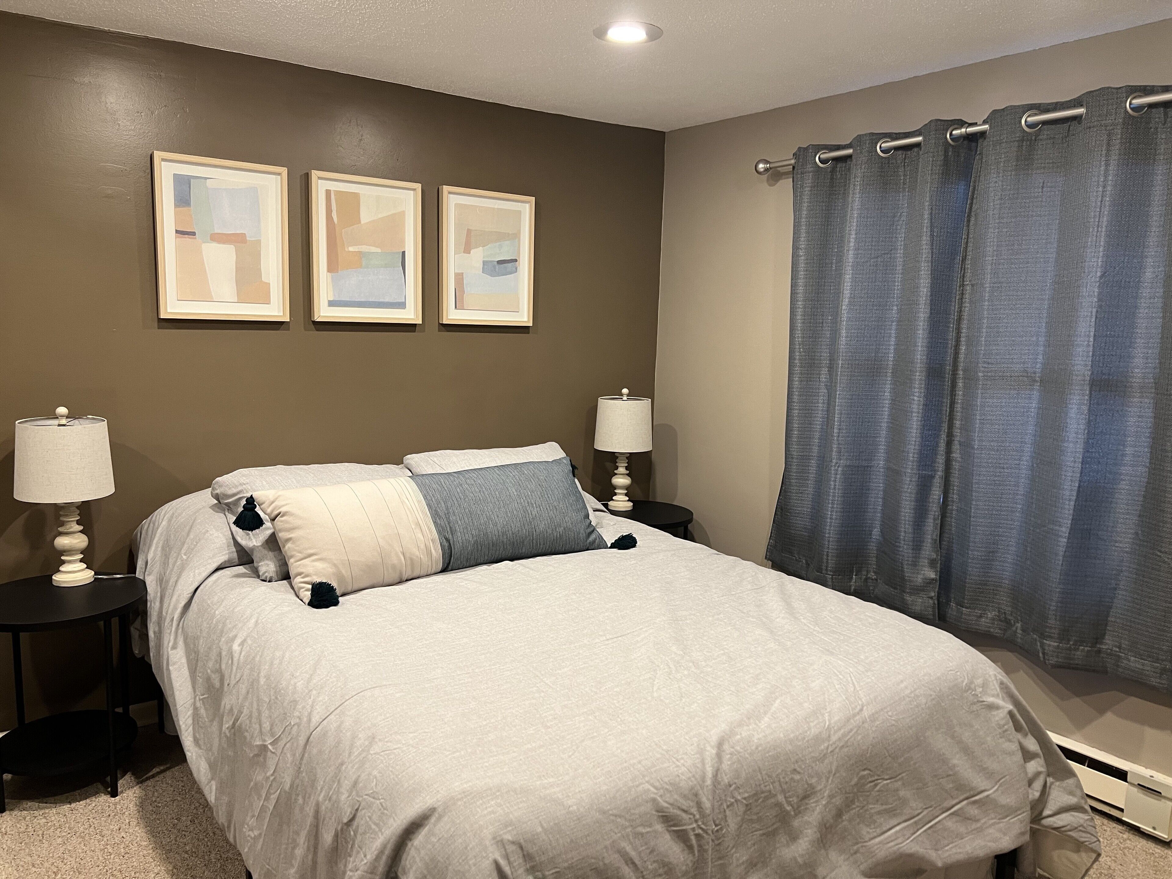 A bedroom with a large bed, two bedside tables with lamps, a window with blue curtains, and three framed abstract paintings on the wall.