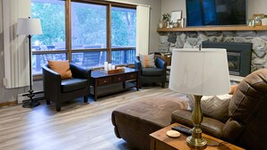 A living room with a fireplace, a TV mounted above it, and a large window providing a view of the outdoors.