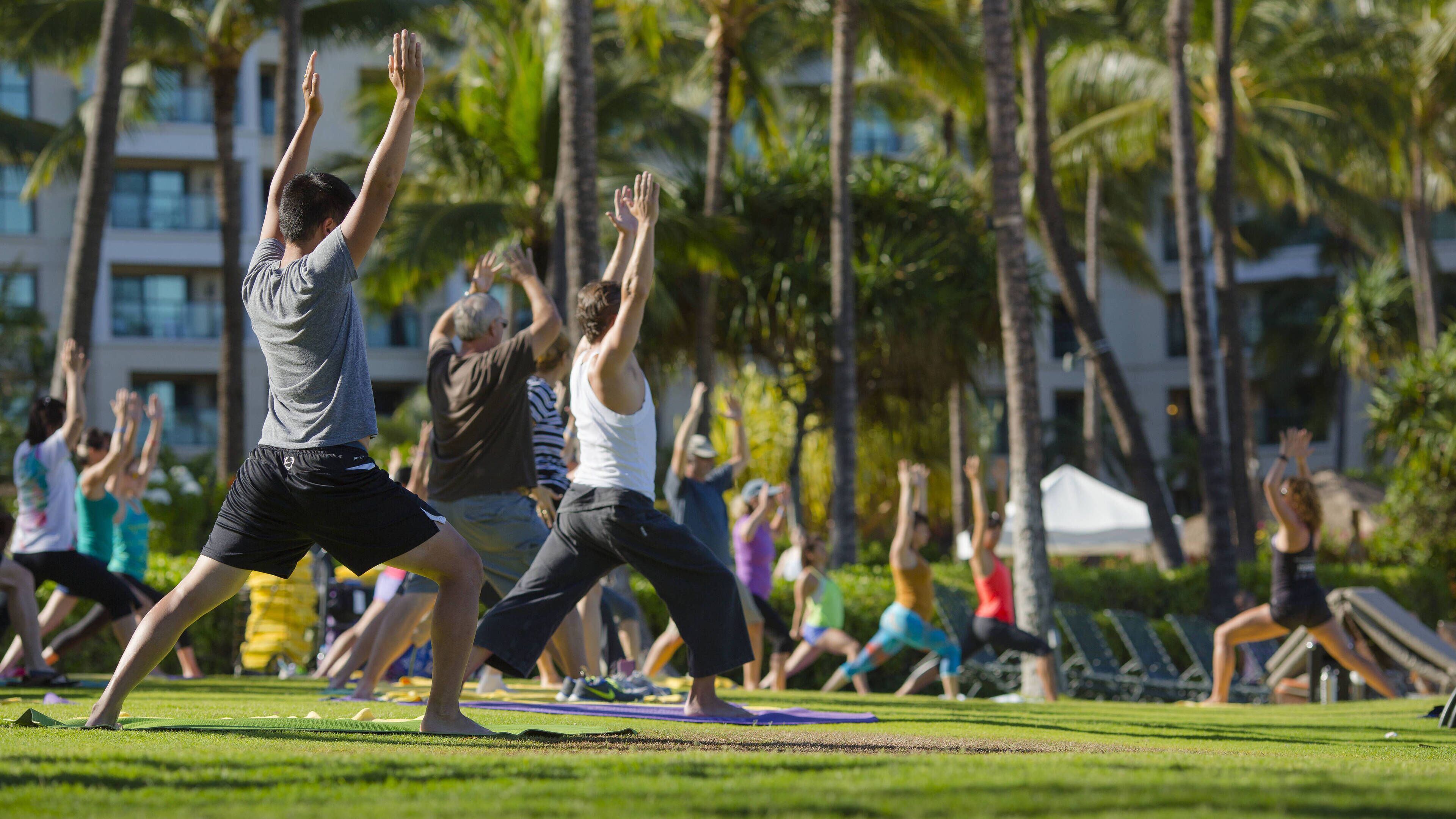 A group of people practicing yoga outdoors on a grassy area with trees and buildings in the background.