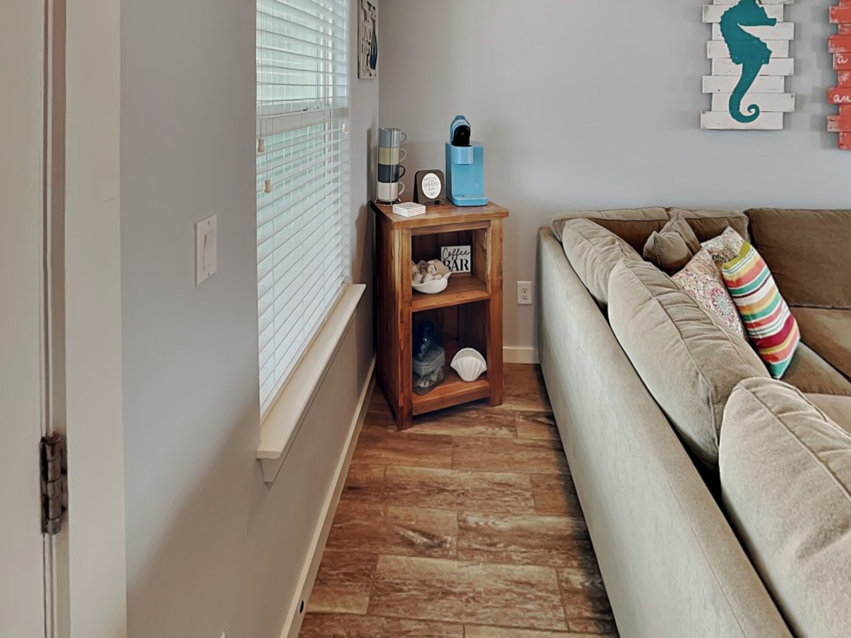 Un salon avec un canapé beige, une table d’appoint en bois sur laquelle se trouve une machine à café, et un mur décoré d’une représentation d’hippocampe.