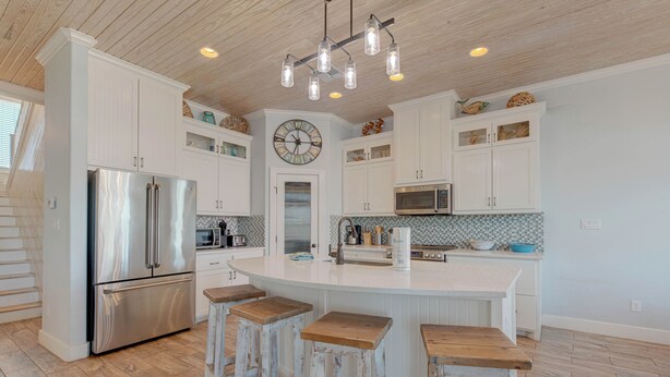 A kitchen with white cabinetry, a stainless steel refrigerator, a microwave, and a large island with stools. - Postcard from Paradise by Silver Sands Vacation Rentals (Port Aransas)