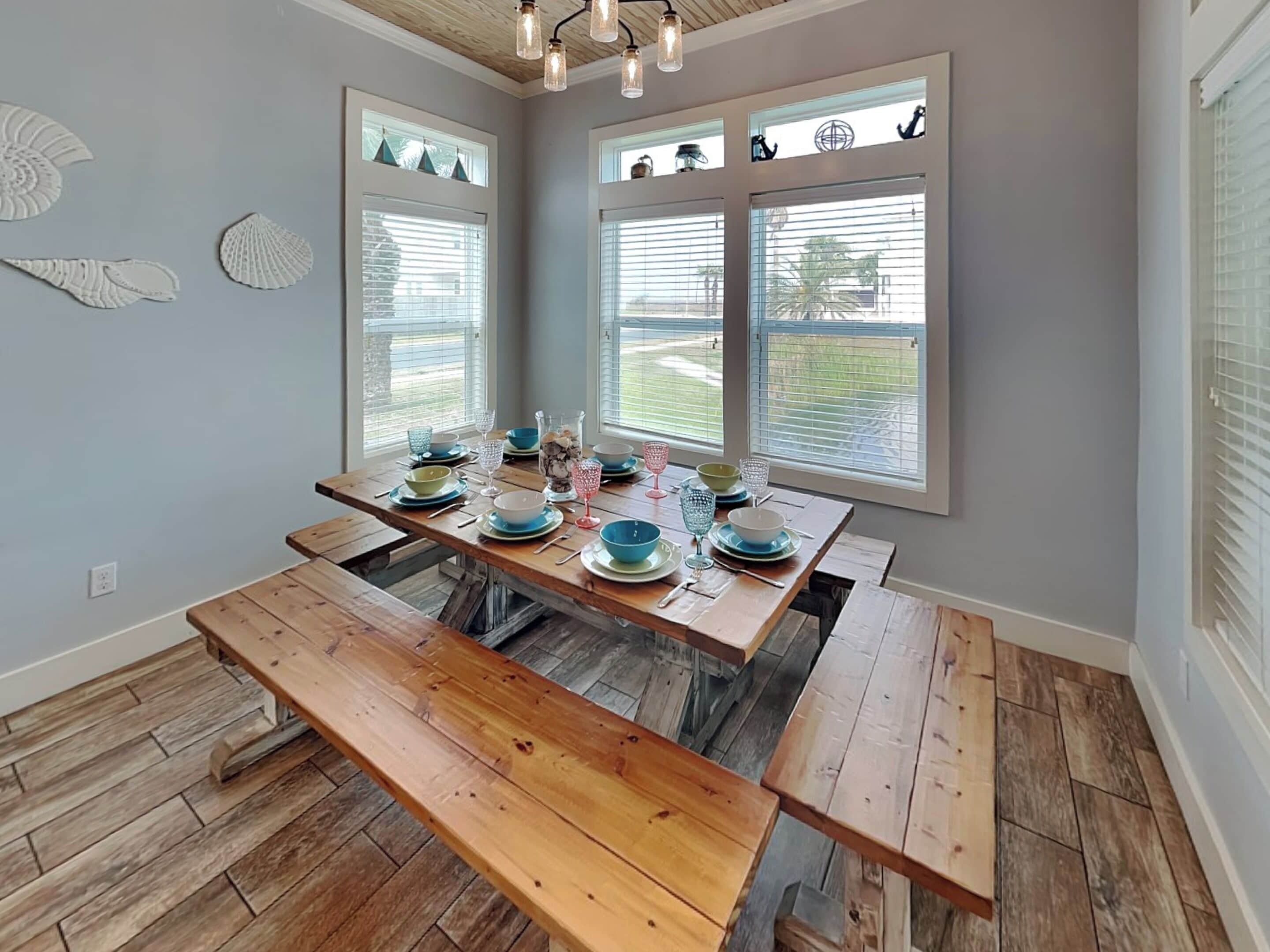 A dining area with a wooden table set for four, large windows with blinds, and a ceiling with exposed beams and hanging lights.