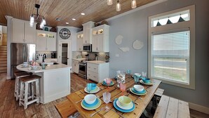 A modern kitchen with white cabinetry, a wooden island, and a dining table set with blue and green plates and glasses. - Postcard from Paradise by Silver Sands Vacation Rentals (Port Aransas)