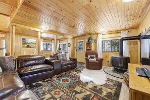 A cozy cabin interior with a leather sofa, a plush armchair, a rug, and a wood-burning stove. - Rustic River Retreat | 2 Cabins, Hot Tub, Fire Pits, Sleeps 24 (Island Park)