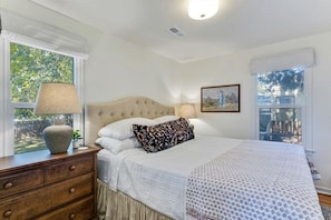 A bedroom with a large bed, a nightstand, a lamp, a window with a view of trees, and a framed picture on the wall.