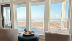 A room with a large window overlooking a beach, two armchairs, and a black ottoman with decorative items. - The Sanctuary by Silver Sands Vacation Rentals (Port Aransas)