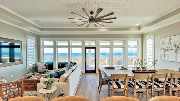 A spacious room with a large window overlooking the beach, a ceiling fan, and a dining area with wooden chairs and a table. - The Sanctuary by Silver Sands Vacation Rentals (Port Aransas)