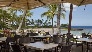 An outdoor dining area with tables and chairs, overlooking a beach and ocean.