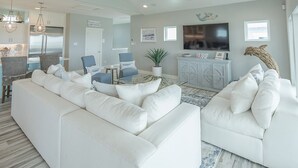 A modern living room with white sofas, a coffee table, and a TV. There is a kitchen area with a refrigerator and a dining table. - All Decked Out by Silver Sands Vacation Rentals (Port Aransas)