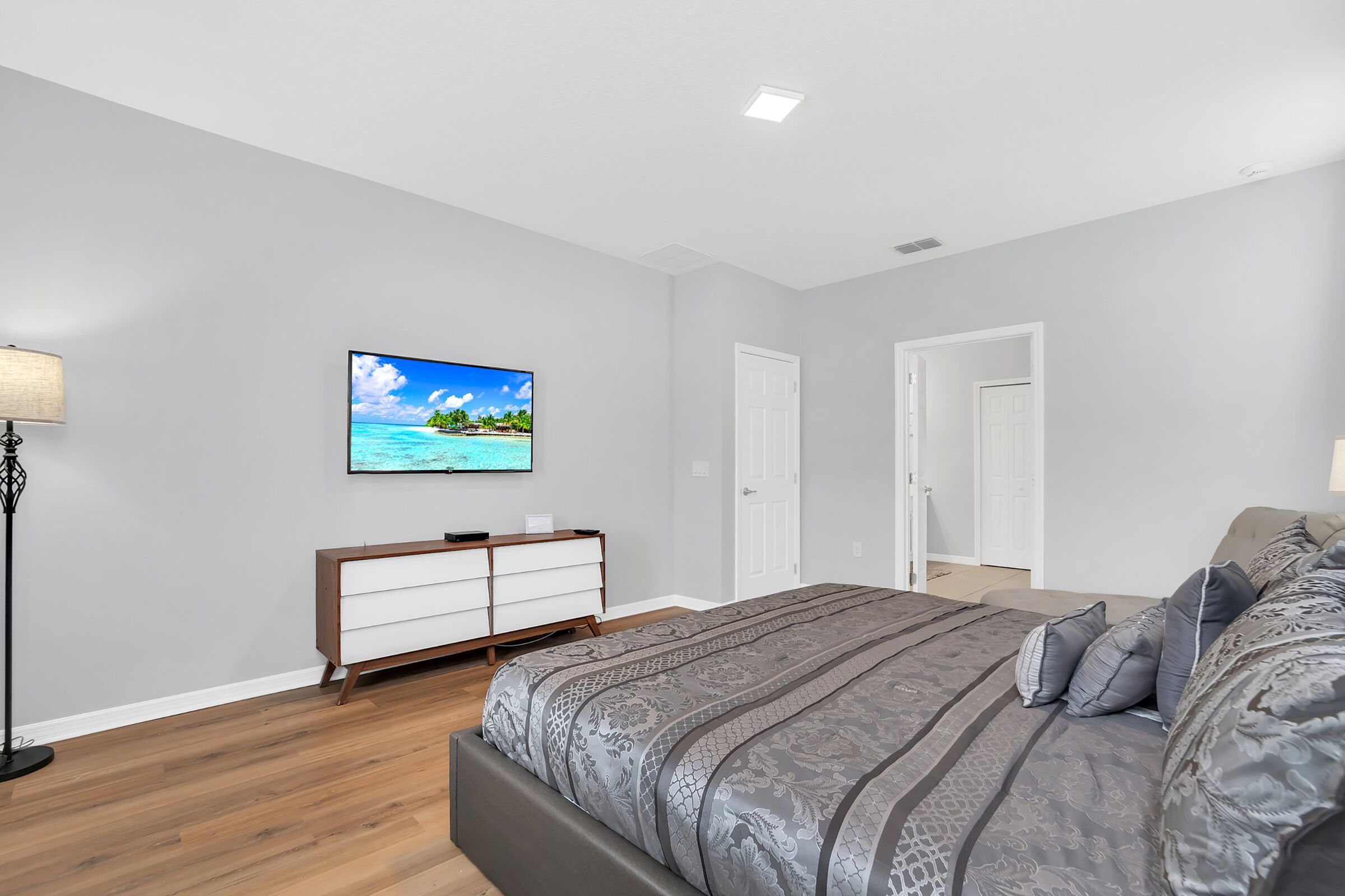 Une chambre à coucher avec un lit, une commode, un téléviseur fixé au mur et une porte.