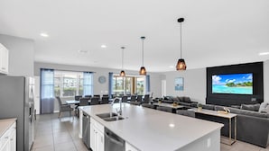 A modern kitchen with a central island, dining area with grey chairs, a sofa, and a TV wall-mounted above a black panel. - Solterra Escape | 14 Bdr, Game Room, Lego Playroom, Theater, Private Pool (Davenport)