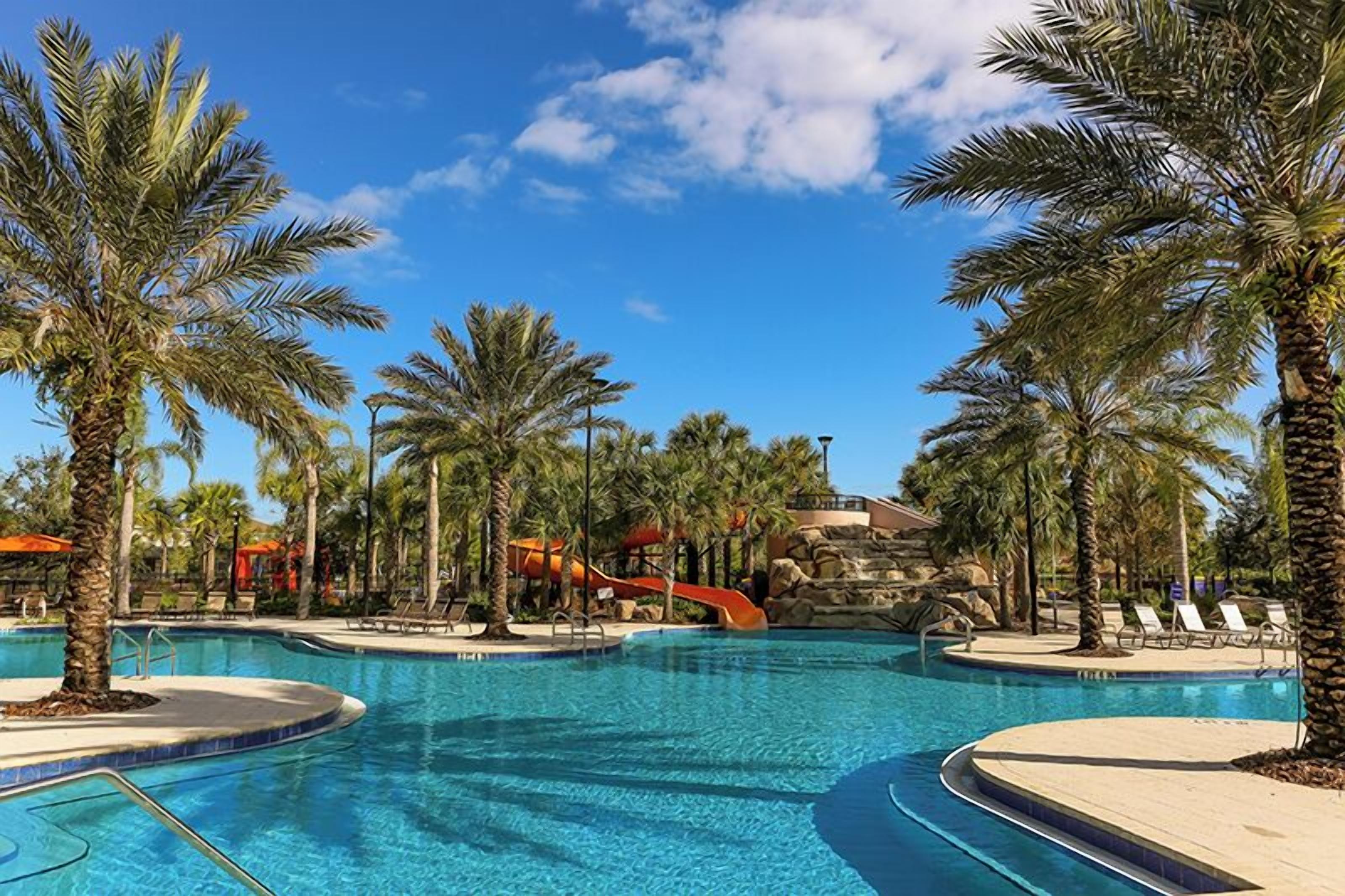 A resort pool area with multiple swimming pools, palm trees, lounge chairs, and a water slide.