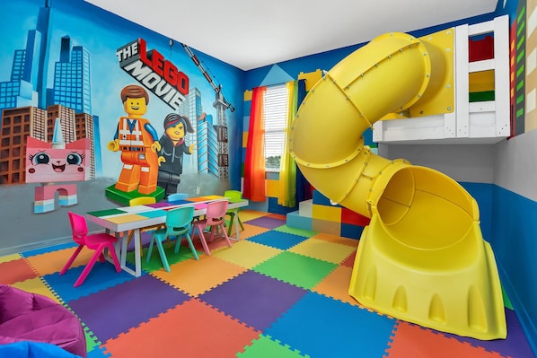 Ein Spielzimmer mit Wandgemälde zu Legostein-Film, bunten Teppichen, einer gelben Rutsche und einem Tisch mit Stühlen.