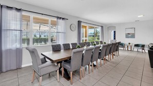 A modern dining area with a long table and grey upholstered chairs, large windows with blinds, and a view of greenery outside. - Solterra Escape | 14 Bdr, Game Room, Lego Playroom, Theater, Private Pool (Davenport)