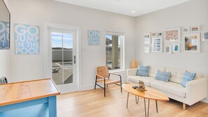 A modern living room with a white sofa, a wooden coffee table, and a wicker chair. There are two signs on the wall, one reading "GO JUMP IN THE POOL" and the other "ON BEACH TIME". - The White House by Silver Sands Vacation Rentals (Port Aransas)