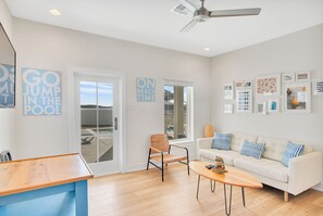 A modern living room with a white sofa, a wooden coffee table, and a wicker chair. There are two signs on the wall, one reading "GO JUMP IN THE POOL" and the other "ON BEACH TIME". - The White House by Silver Sands Vacation Rentals (Port Aransas)