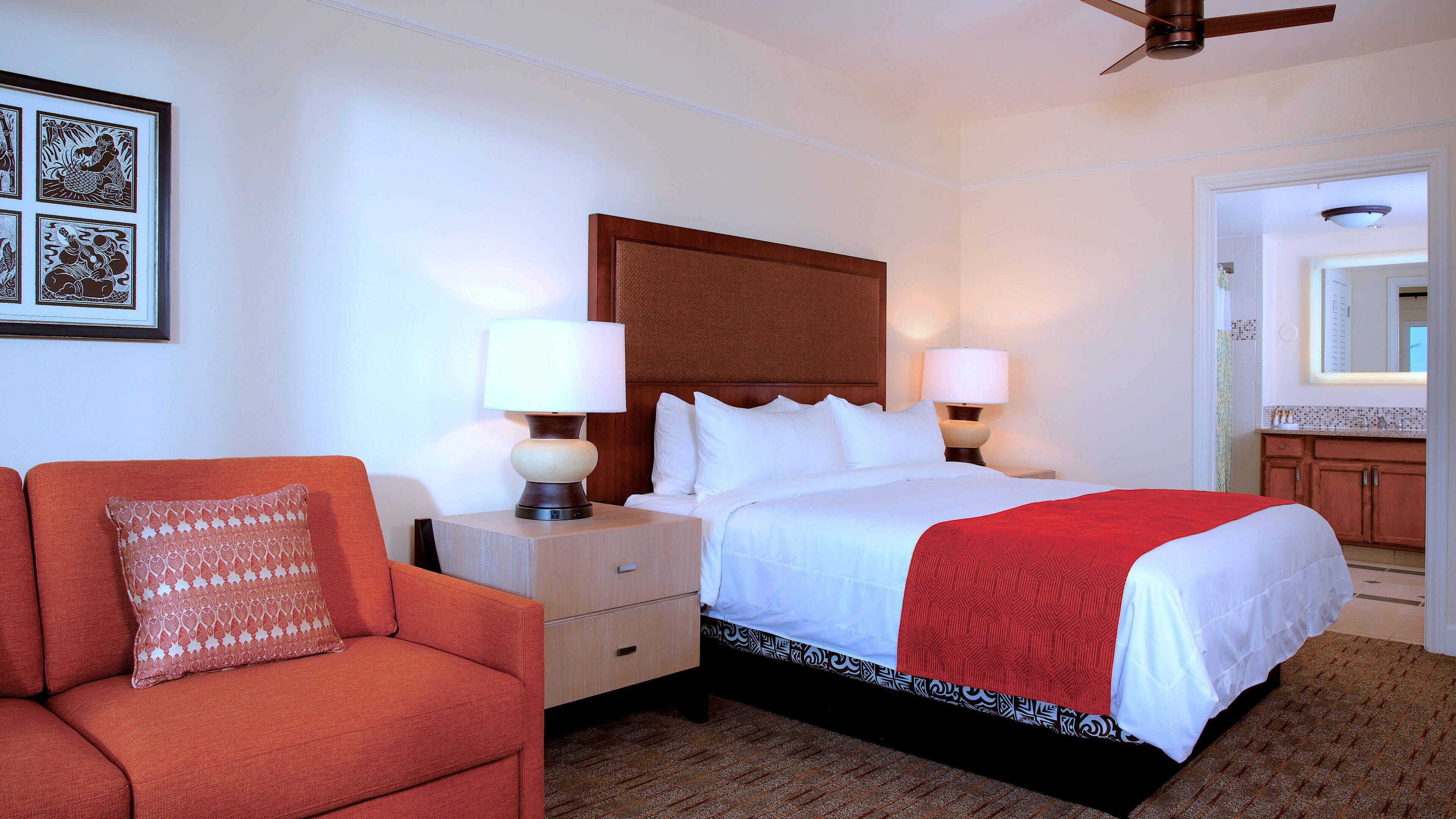 Quarto de hotel com cama, duas mesas de cabeceira com luminárias, uma poltrona vermelha e um ventilador de teto.