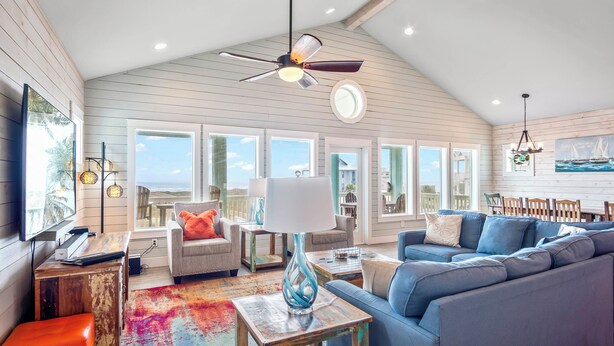 A spacious living room with a high ceiling, large windows, a ceiling fan, and a colorful rug. - Beach-A-Holic by Silver Sands Vacation Rentals (Port Aransas)