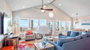 A spacious living room with a high ceiling, large windows, a ceiling fan, and a colorful rug.