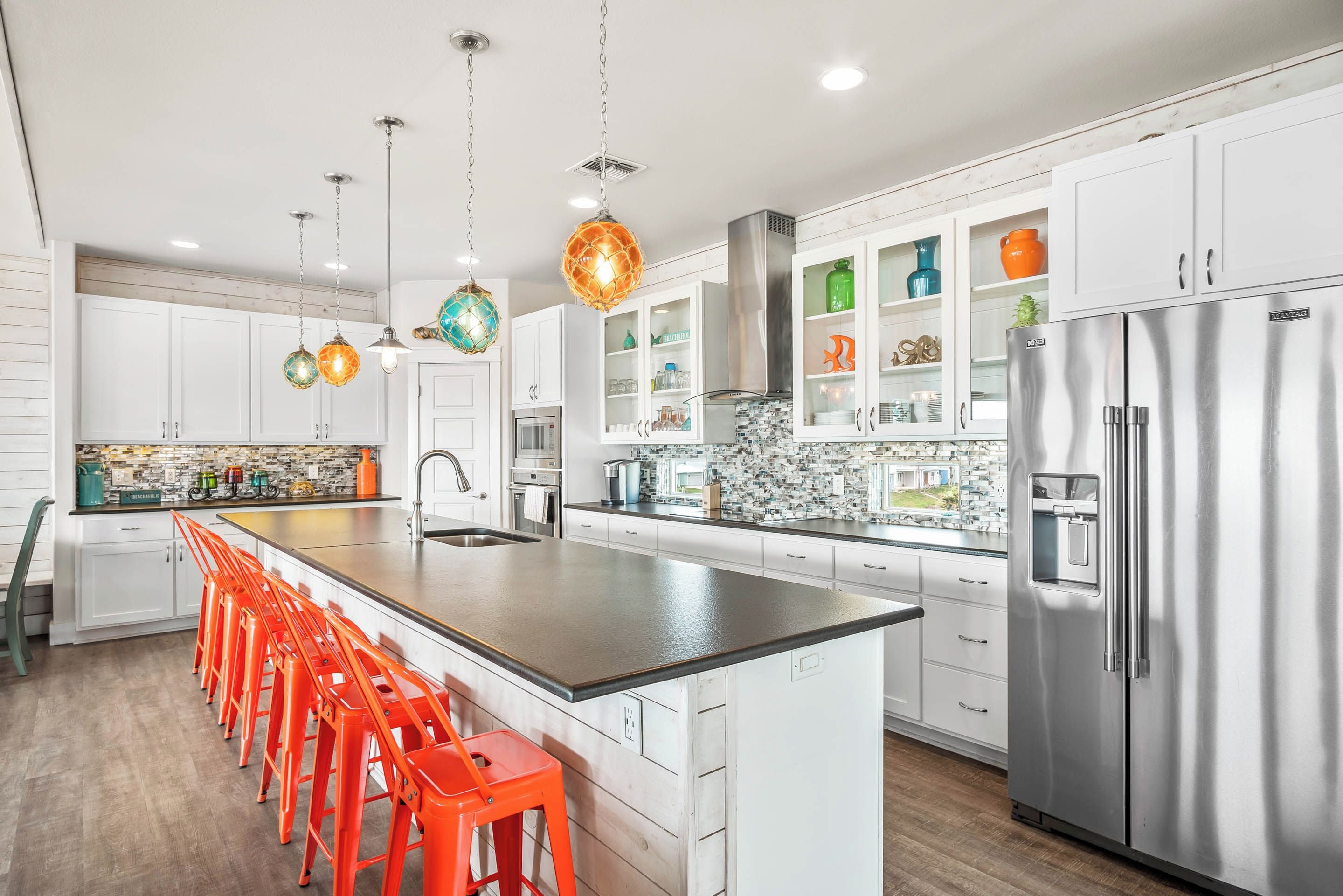 A modern kitchen with white cabinets, a stainless steel refrigerator, and a long island with orange stools.