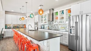 A modern kitchen with white cabinets, a stainless steel refrigerator, and a long island with orange stools. - Beach-A-Holic by Silver Sands Vacation Rentals (Port Aransas)